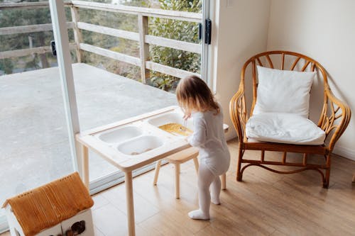 Back view of unrecognizable little girl in casual white clothes standing at table near window and playing with sensory elements in light room at home
