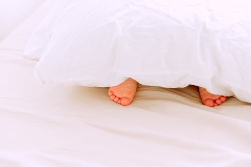 Free Crop tiny feet of little child lying under white blanket on comfortable bed in light bedroom at home Stock Photo