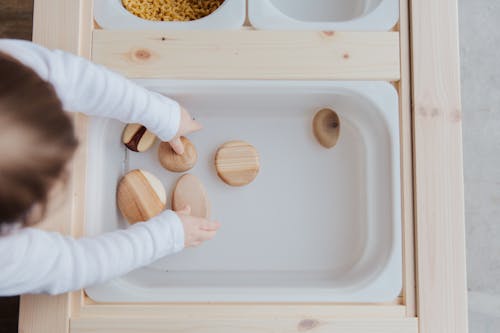 Free From above view of unrecognizable little child playing with wooden round stones in white plastic container at wooden table at home Stock Photo