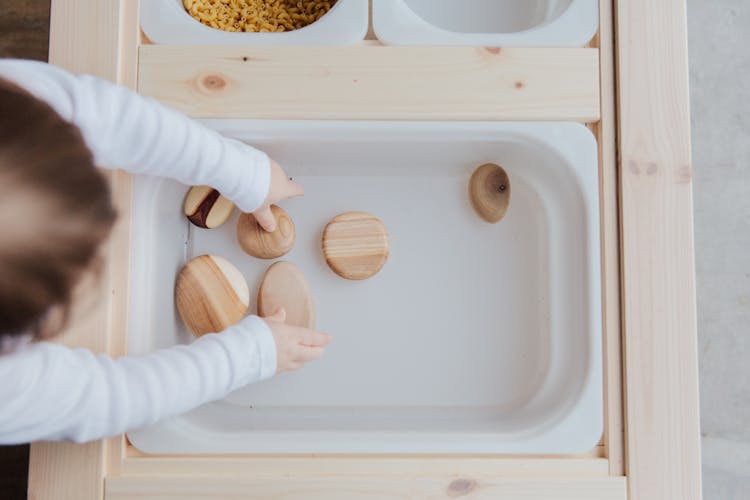 Unrecognizable Kid Playing With Stones In White Container At Table At Home