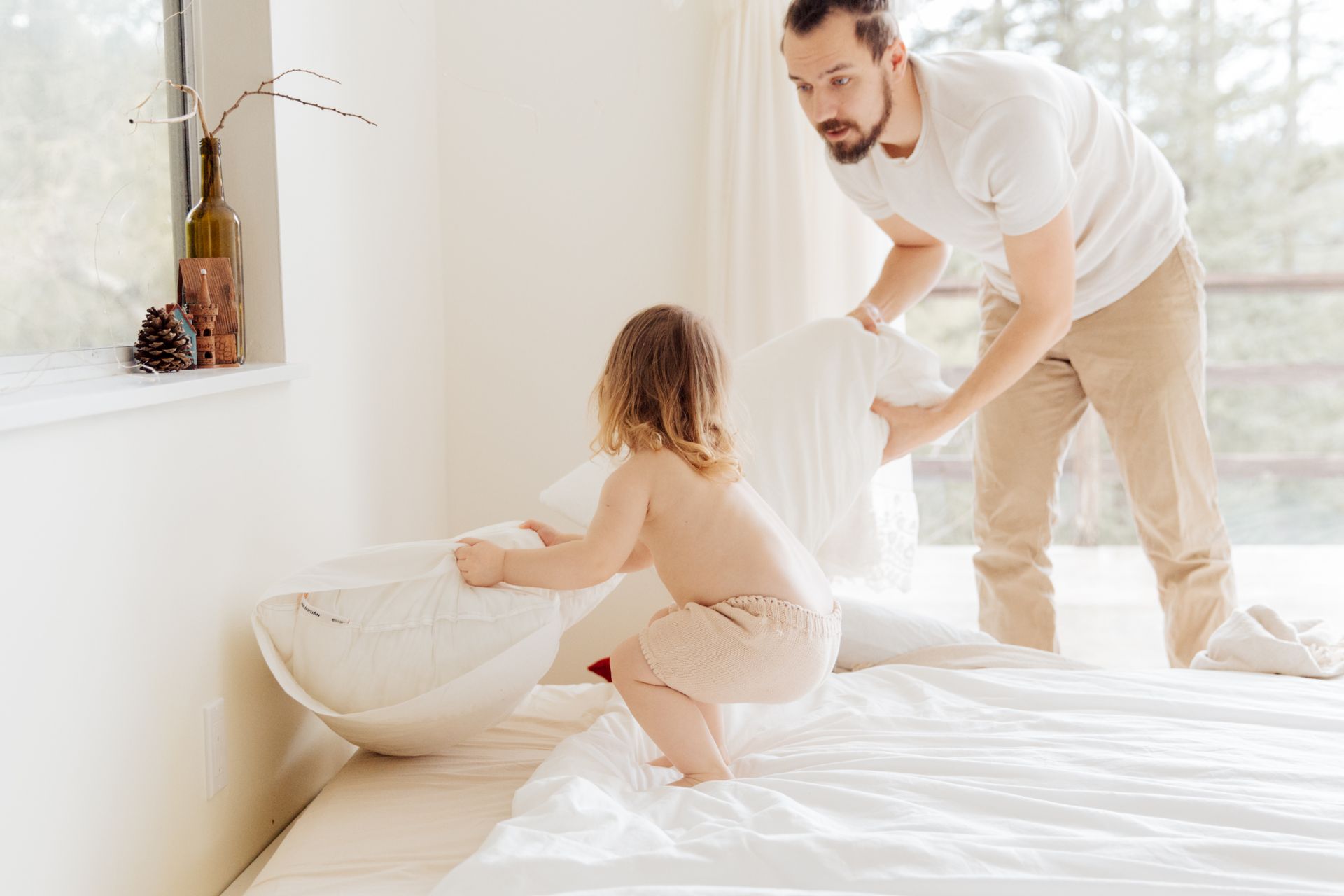 Father and child enjoying a playful pillow fight in a bright, cozy bedroom setting.