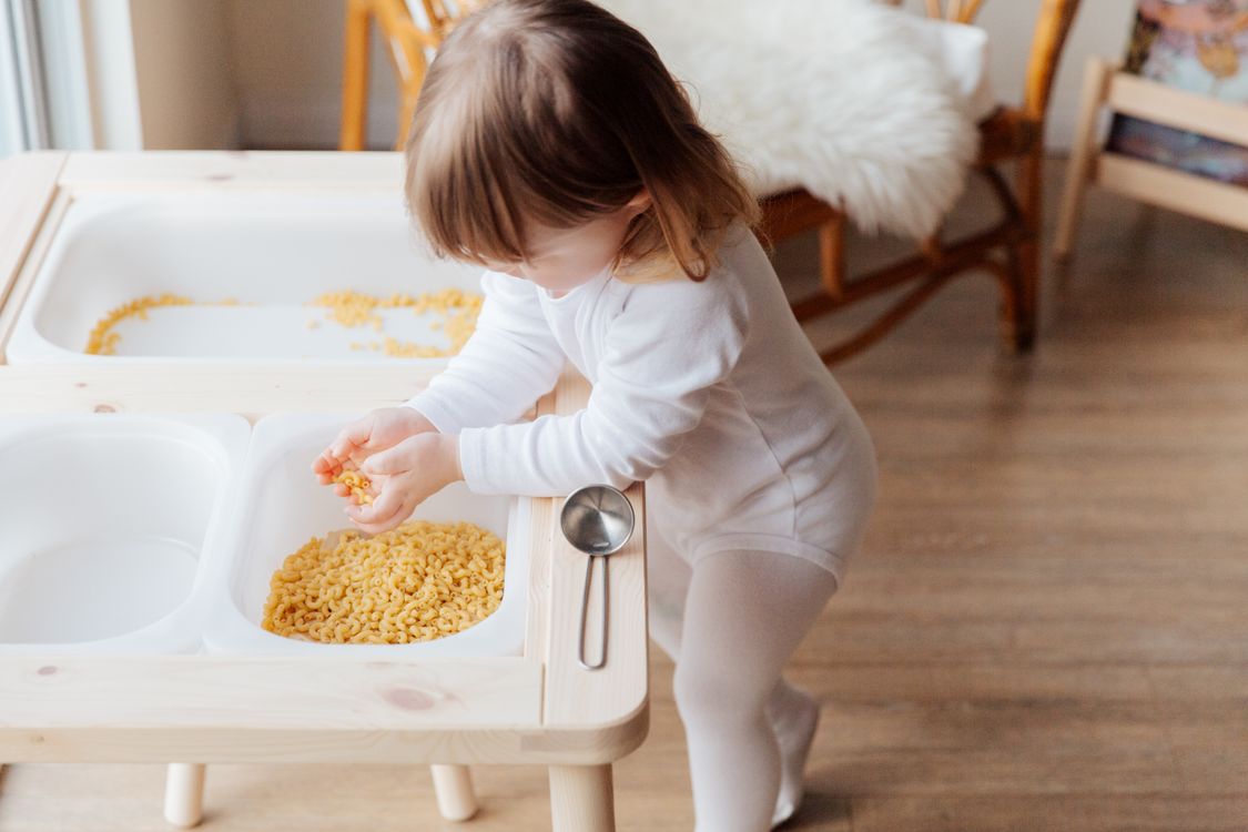 A child's hand holds a piece of macaroni pasta during a sensory play activity.