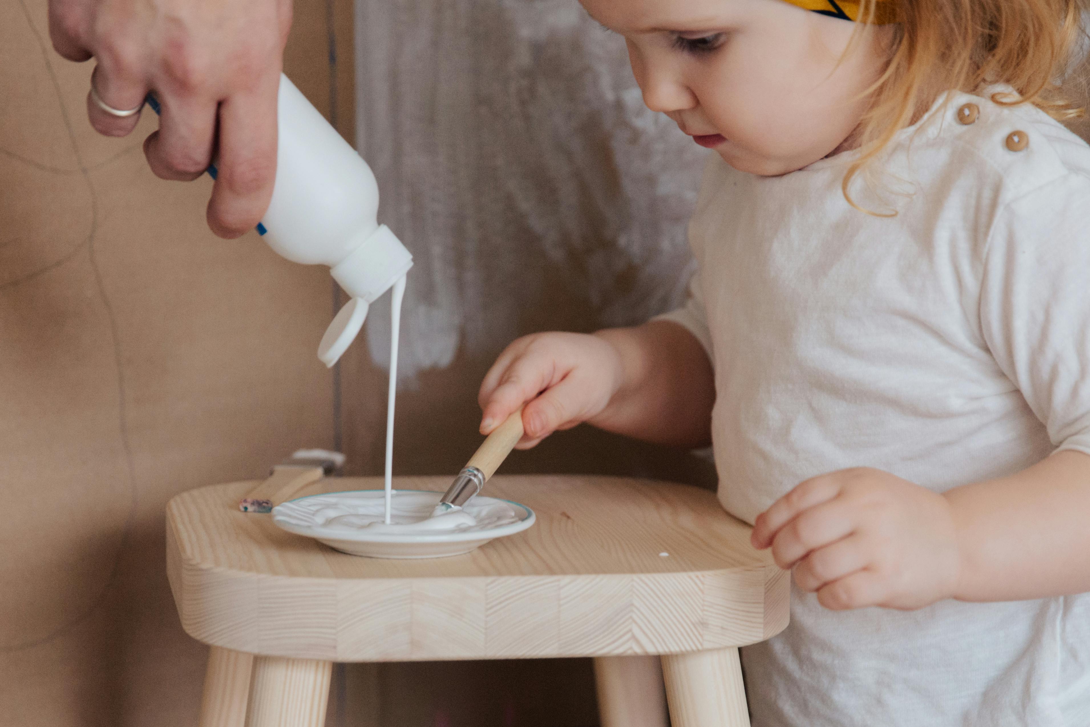 anonymous cute toddler girl holding brush for getting white paint from plate standing on chair