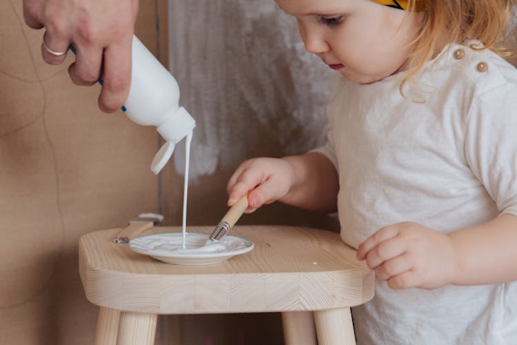 Anonymous Cute Toddler Girl Holding Brush For Getting White Paint From Plate Standing On Chair