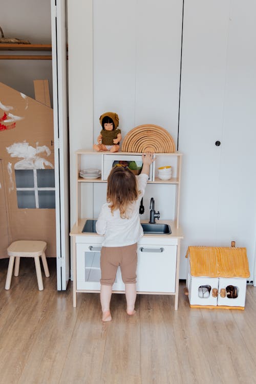 Back view of adorable cute toddler girl playing with toy kitchen while standing barefoot on floor of playroom at home