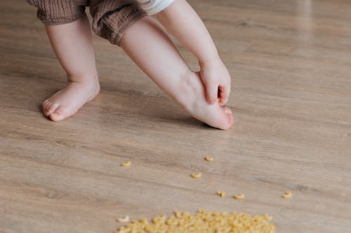 Free Crop faceless toddler standing barefoot on floor and trying to remove stuck pasta from foot while playing and developing fine motor skills at home Stock Photo