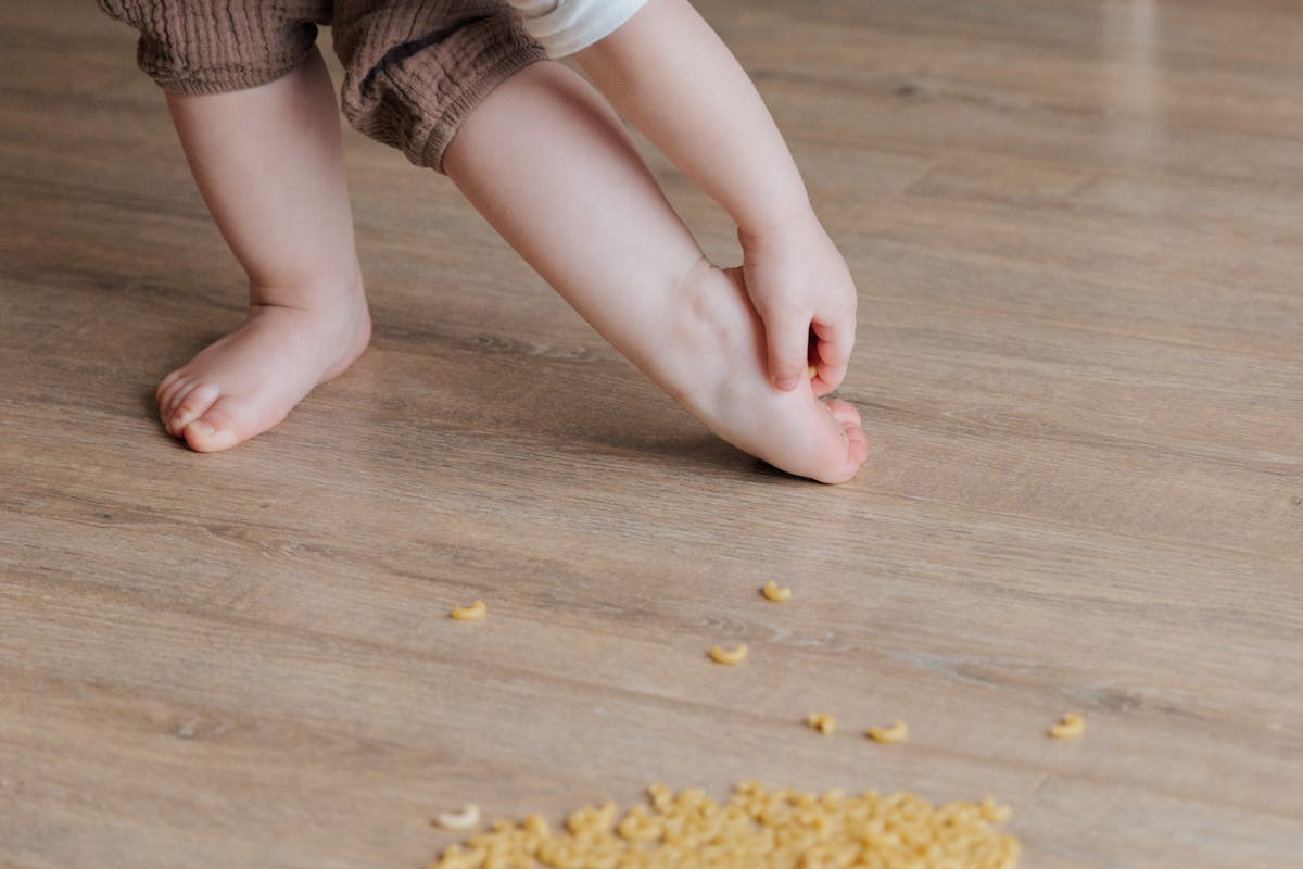 Crop faceless toddler standing barefoot on floor and trying to remove stuck pasta from foot while playing and developing fine motor skills at home