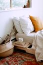 Stylish cozy bedroom interior with bed located near window with white linen, cushions,  tray with cup of coffee, basket with dry wooden branches and carpet on floor