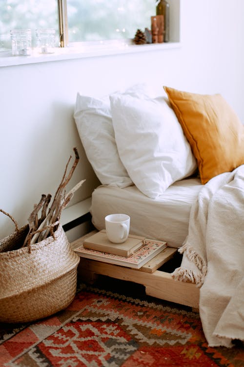 Stylish cozy bedroom interior with bed located near window with white linen, cushions,  tray with cup of coffee, basket with dry wooden branches and carpet on floor