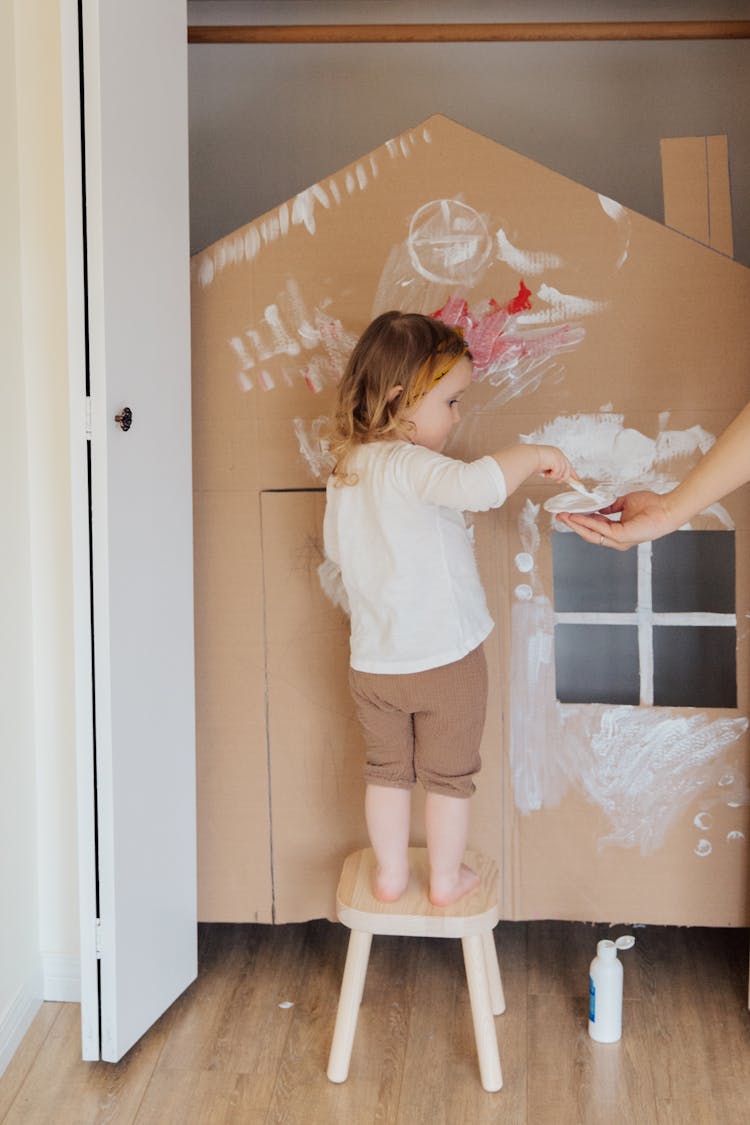 Girl In White Shirt And Brown Shorts Standing In Front Of Cardboard Play House