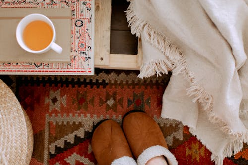 From above of cozy bedroom interior with white plaid, brown warm slippers on carpet, wicker basket and cup of tea on tray