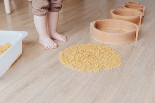 Crop faceless toddler standing barefoot on floor and playing with round wooden shapes of different size and pasta developing fine motor skills at home