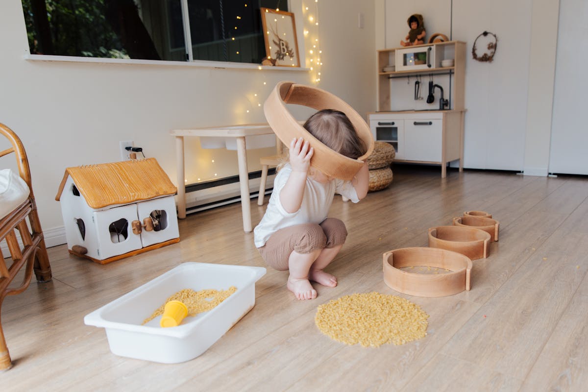 Full length of anonymous toddler squatting barefoot on floor playing with round wooden shapes of different size and pasta and putting biggest shape on while developing fine motor skills at home