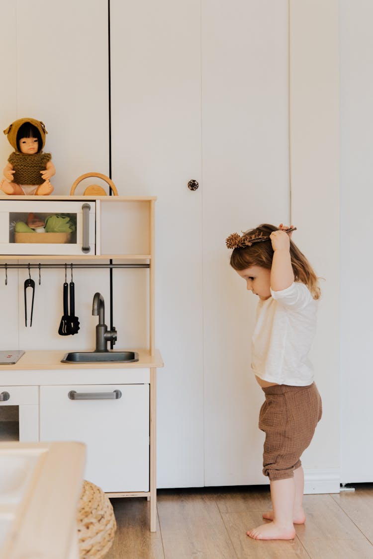 Cute Toddler Girl Trying To Put Christmas Wreath On Head While Standing In Playroom