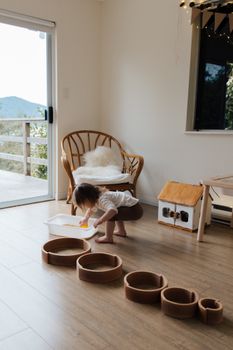 一名无脸幼儿赤脚站在地板上，正用白色容器玩着不同大小的圆形木制形状，这是他在家中发展精细运动技能时的后视全身照