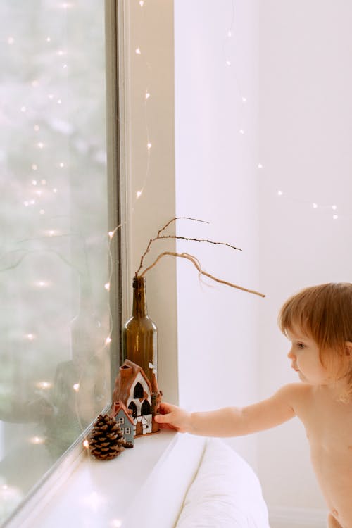 Toddler girl touching bottle with dry branches on windowsill