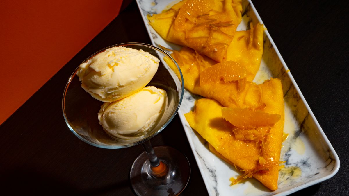 Folded crepes glazed with orange sauce and segments beside a glass of vanilla ice cream scoops