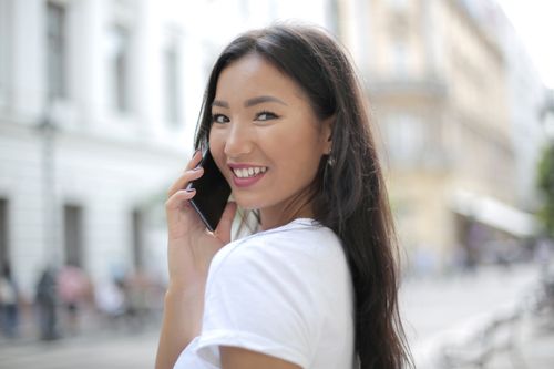 Free Asian woman smiling while using a smartphone outdoors on a sunny day. Casual and joyful interaction. Stock Photo