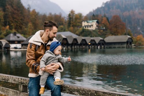 Free Father holds child by a scenic lake with autumn foliage, enjoying outdoor family time. Stock Photo