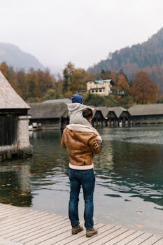 A father carrying his child at a scenic lakeside with mountains in the background during autumn.