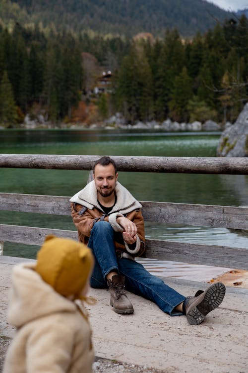Calm young bearded father in warm clothes sitting on wooden pier and leaning on railing while looking at little child