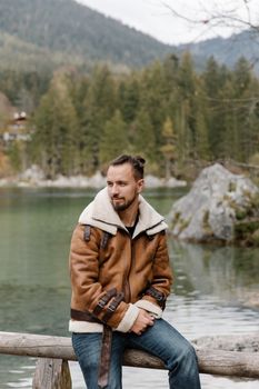 一位英俊的男人穿着棕色夹克在湖边摆姿势，背景是风景秀丽的山峦