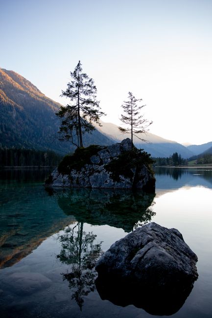 Serene lake scene with rocky island and tree reflections against mountains at sunset.