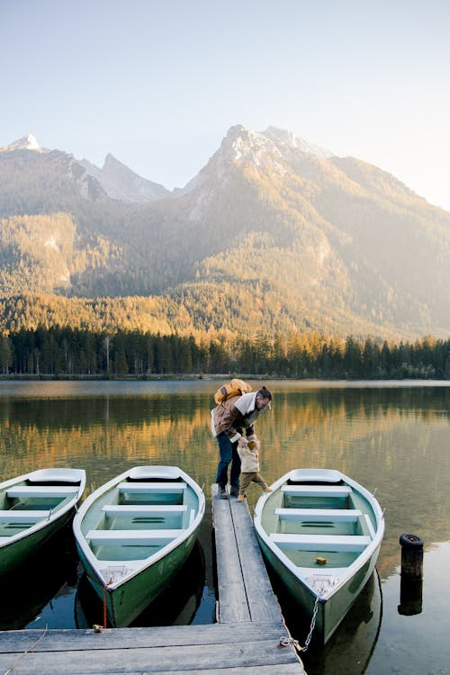 Father with little kid in warm clothes standing on wooden pier near boats on lake shore in mountains and enjoying sunny day in nature
