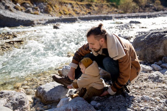 Bearded young father with little child in casual warm clothes sitting on rocky ground near river and enjoying time together while weekend