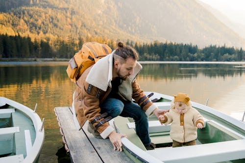 Bearded father with backpack in casual warm outerwear standing on wooden pier and holding hand of little kid standing in boat on lake in mountains