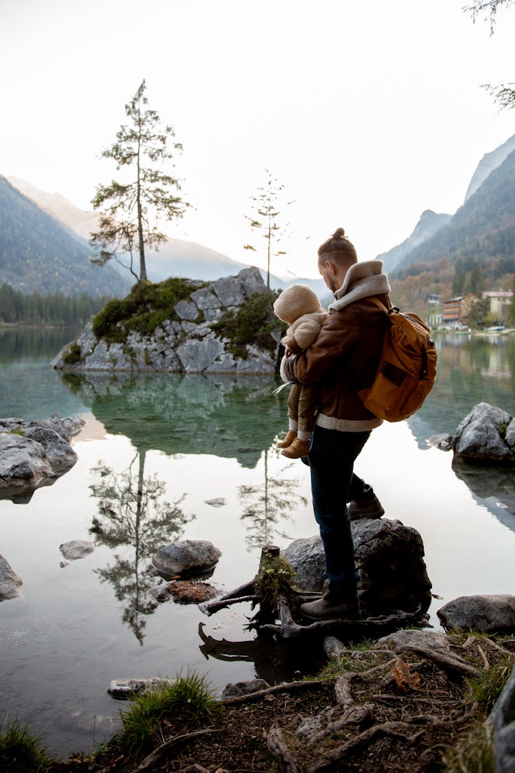 Father With Little Kid On Hands Standing On Stone On Lake Shore In Forest