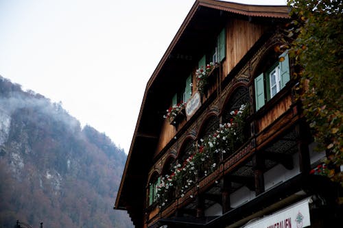 Facade of traditional wooden house decorated with colorful potted flowers located in quiet valley with forested mountains in cloudy weather