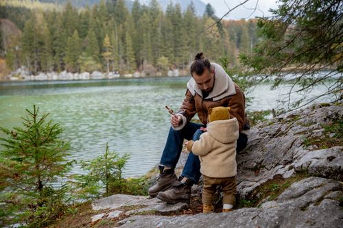 Free Father and little kid in casual warm clothes resting on rocky ground near lake in calm forest on overcast day in autumn Stock Photo