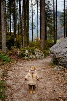 一个穿着米色夹克的可爱幼儿，走在被高大树木包围的森林小路上