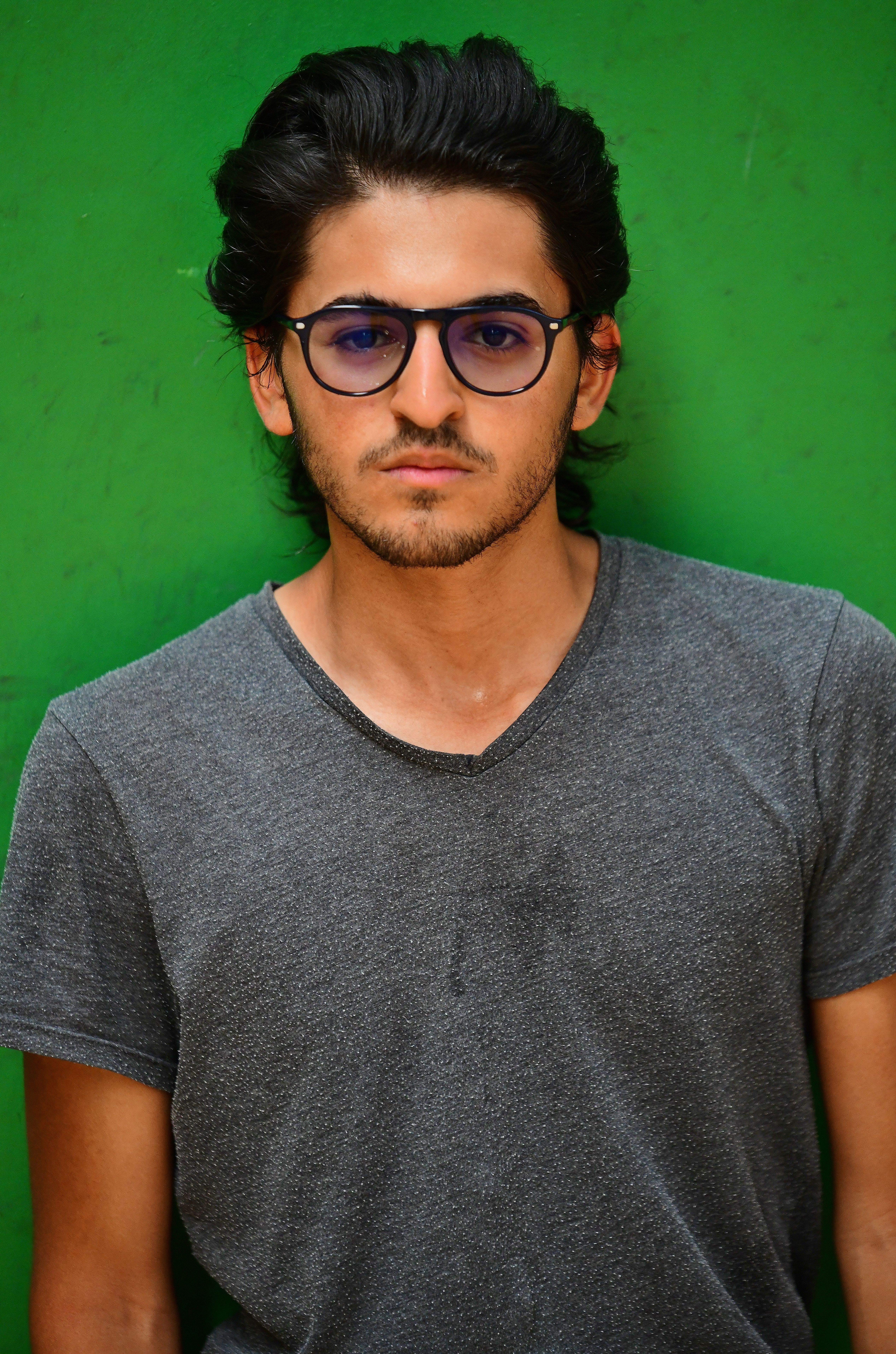 Young man wearing glasses and gray t-shirt, standing against a green wall.