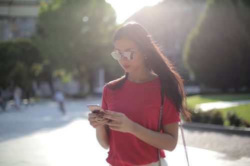 Free Asian woman in red t-shirt texts outdoors in bright daylight with sunglasses. Stock Photo