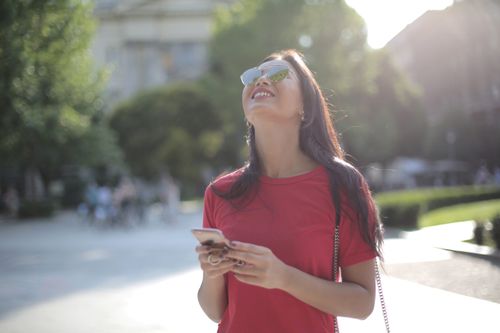 Free Smiling young woman in red t-shirt and sunglasses enjoying a sunny day outdoors with her smartphone. Stock Photo