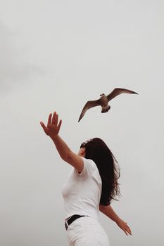 A woman in white reaches out to a soaring bird under a cloudy sky, expressing freedom and connection with nature.