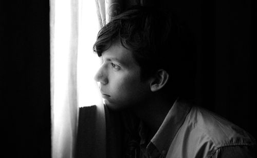 Free Black and white portrait of a young man looking out a window, deep in thought. Stock Photo