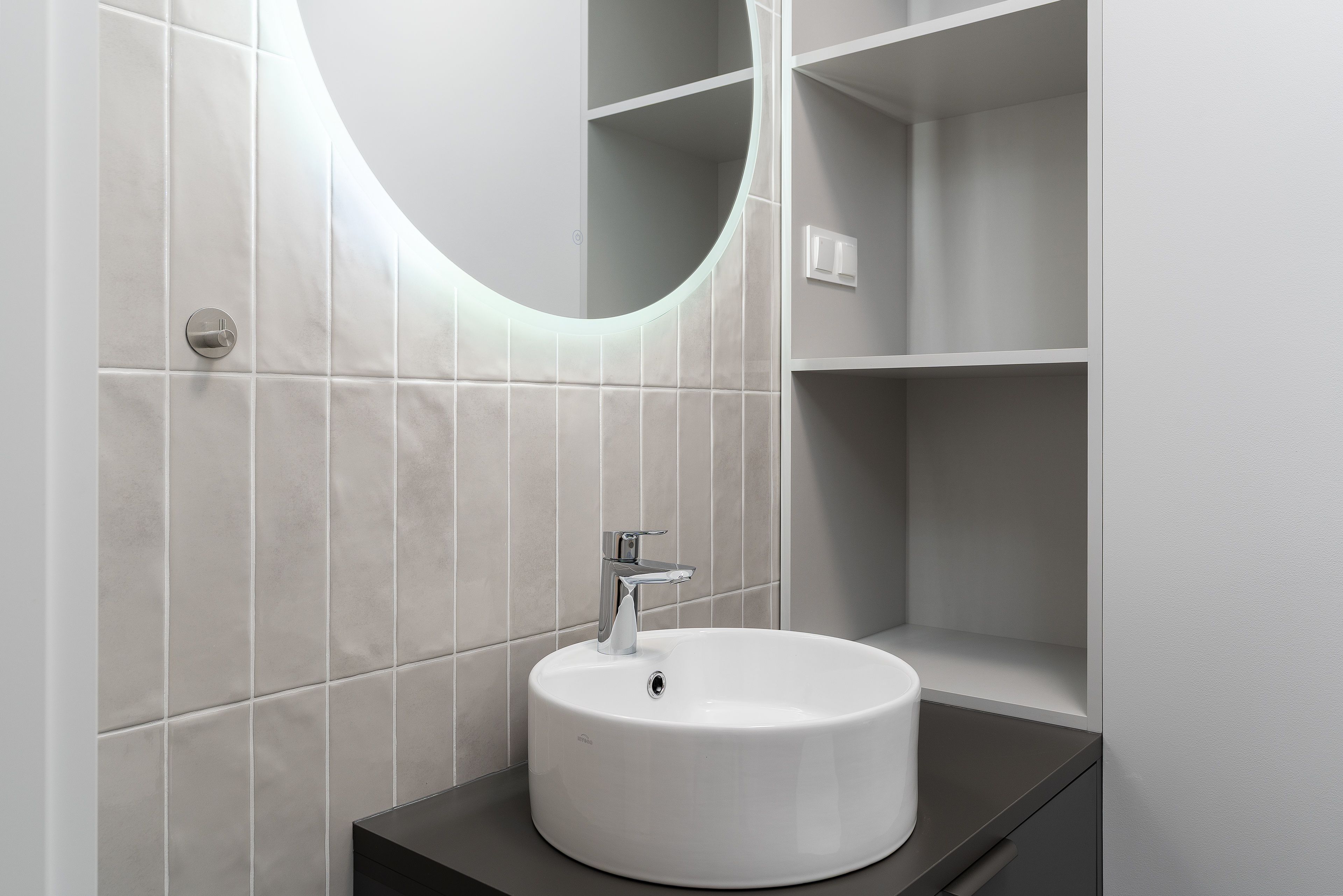 Modern bathroom sink with round mirror, gray tile, and built-in wall shelves