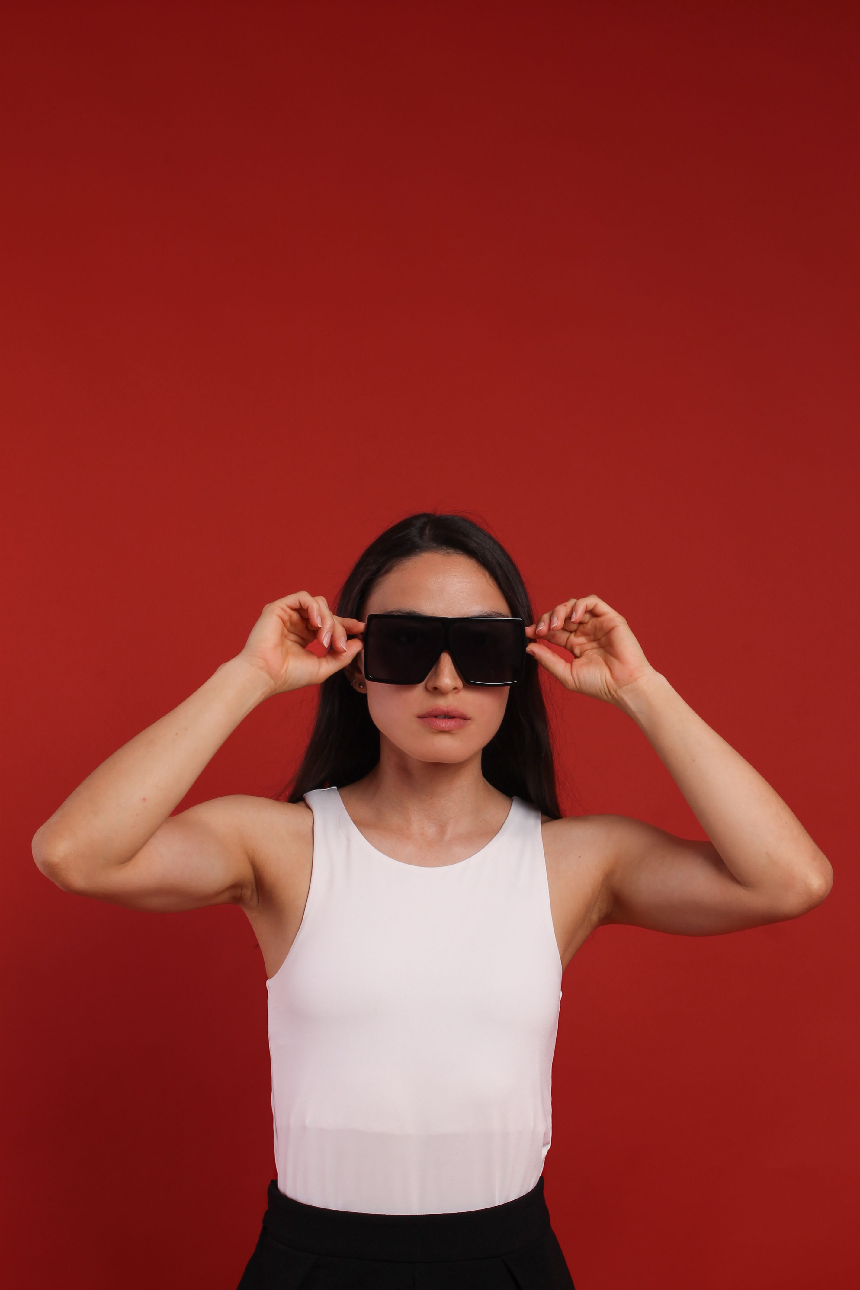 Woman in white top and sunglasses posed against vibrant red backdrop, exuding modern style.