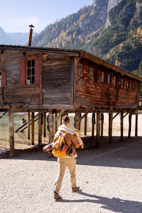 Free Back view of unrecognizable man with tourist equipment and yellow backpack walking along old wooden house placed by river near mountains Stock Photo