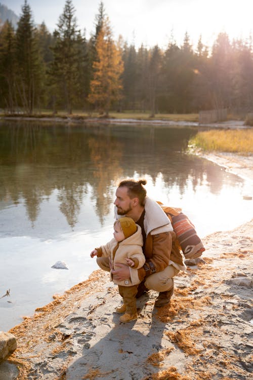 Free Man in Brown Jacket Sitting on Rock Near Lake Stock Photo