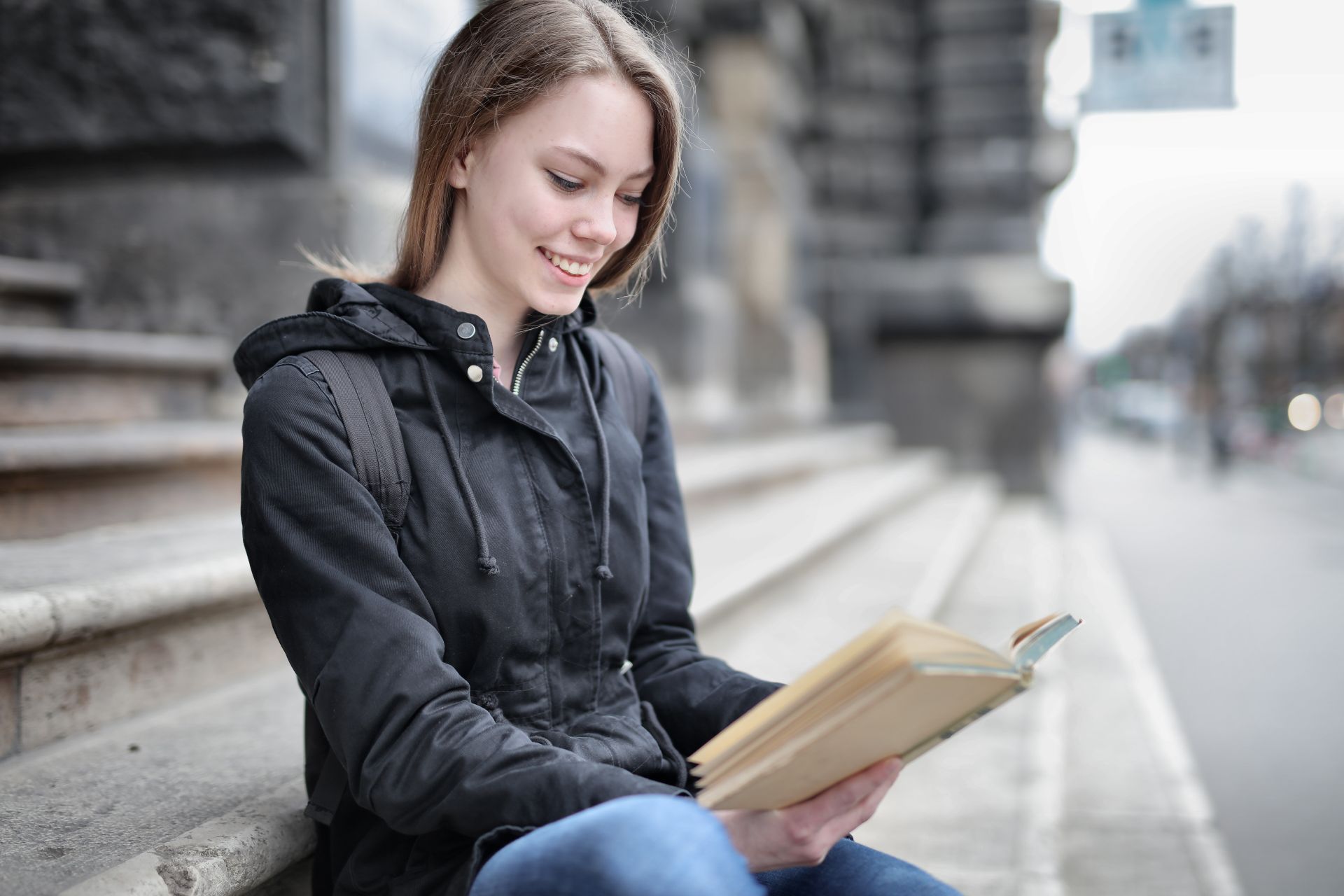 A young woman in casual attire reading a book while sitting on city steps, smiling.