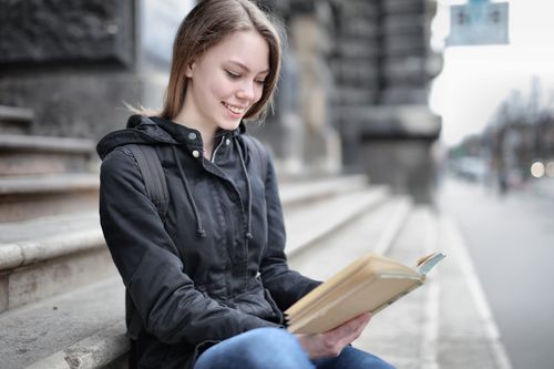 Free A young woman in casual attire reading a book while sitting on city steps, smiling. Stock Photo