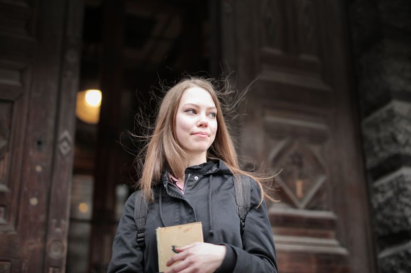 Portrait of a young woman holding a book outside by a wooden door.