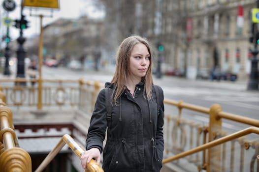 A young woman in a black coat exits a subway in an urban street scene.