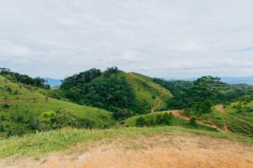 Free Scenic view of lush green hills and a winding dirt trail under a cloudy sky. Stock Photo
