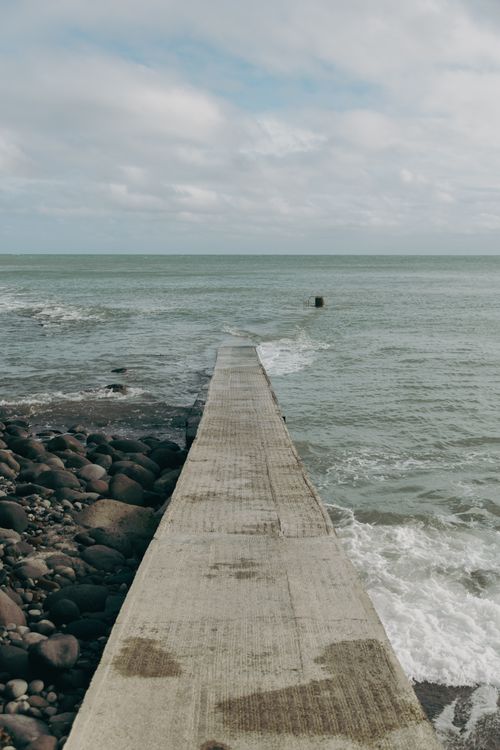 Free A serene view of a concrete pier leading into the vast ocean under a cloudy sky. Stock Photo