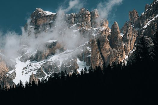 Majestic Dolomite mountains with snowy peaks and mist in a dark forest setting.